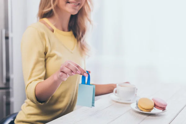 Vue recadrée de la femme tenant un petit sac à provisions près de la tasse et des macarons — Photo de stock