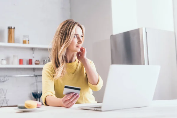 Atractiva mujer rubia sosteniendo la tarjeta de crédito cerca del ordenador portátil en la cocina - foto de stock