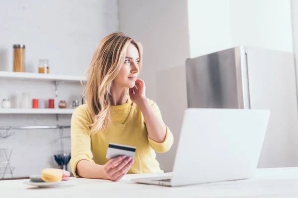 Attractive blonde woman holding credit card near laptop in kitchen — Stock Photo