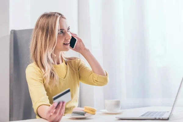 Attractive blonde woman talking on smartphone while holding credit card near laptop — Stock Photo