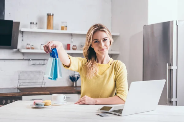 Mujer rubia sosteniendo pequeñas bolsas de compras mientras está sentado en la cocina - foto de stock