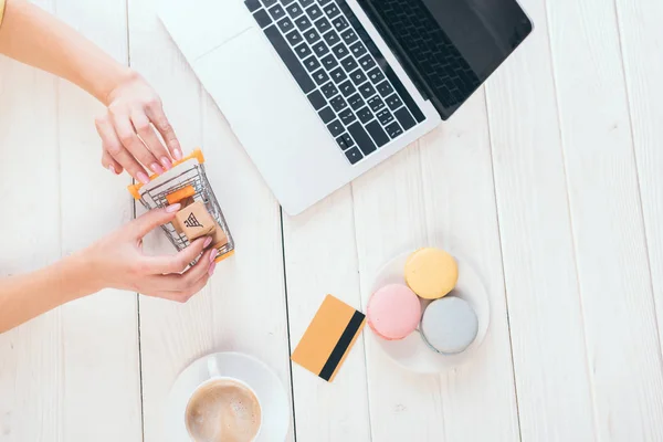 Donna vista dall'alto con carrello della spesa giocattolo con piccole scatole di carta vicino a laptop, tazza e macaron — Foto stock