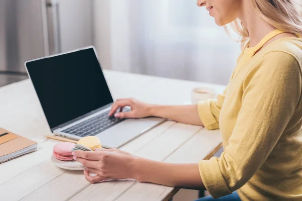 Vista recortada de la mujer escribiendo en el ordenador portátil y la celebración de macarrones - foto de stock