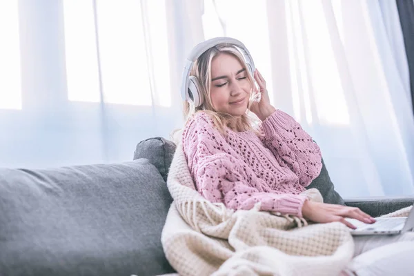 Mujer rubia sonriente escuchando música en auriculares mientras está sentada en casa - foto de stock