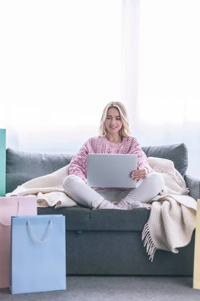 Heureuse femme blonde assise sur un canapé avec ordinateur portable près des sacs à provisions — Photo de stock