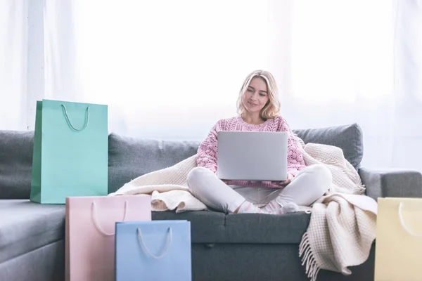 Mujer rubia alegre sentada en el sofá con el ordenador portátil cerca de las bolsas de compras - foto de stock