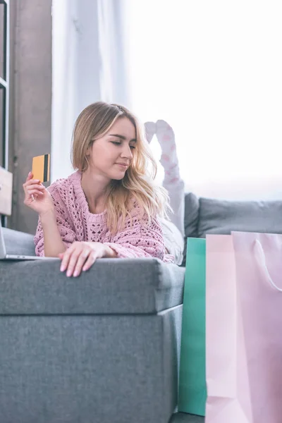 Femme heureuse tenant la carte de crédit tout en étant allongé sur le canapé et en regardant des sacs à provisions — Photo de stock