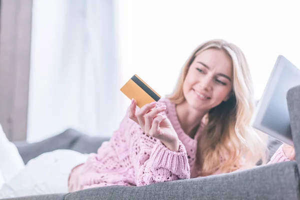 Mujer sonriente mirando a la celebración de la tarjeta de crédito mientras está acostado en el sofá con la tableta digital - foto de stock