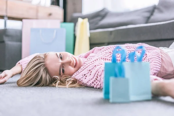 Fröhliche Frau schaut auf Einkaufstüten, während sie zu Hause auf dem Boden liegt — Stockfoto