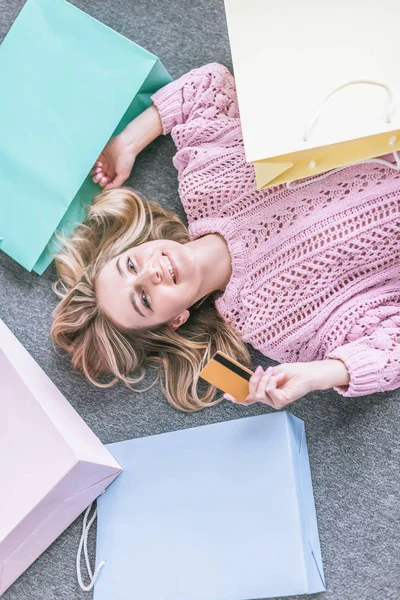 Top view of cheerful woman holding credit card and lying on floor near shopping bags — Stock Photo