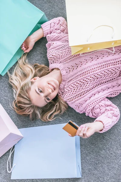 Vista superior da mulher alegre olhando para o cartão de crédito e deitado no chão perto de sacos de compras — Fotografia de Stock