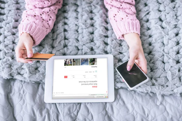 Vista recortada de la mujer que sostiene la tarjeta de crédito y el teléfono inteligente cerca de la tableta digital con aplicación airbnb en la pantalla - foto de stock