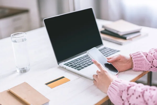 Vista cortada de mulher segurando smartphone perto de laptop com tela em branco — Fotografia de Stock