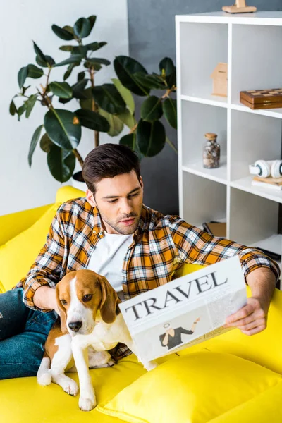 Удивленный молодой человек читает туристическую газету и сидит на диване с симпатичной собакой — стоковое фото