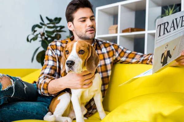 Schöner junger Mann liest Reisezeitung, während er mit Beagle-Hund auf dem Sofa sitzt — Stockfoto