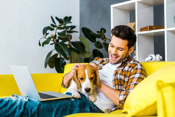 Glücklicher junger Mann mit Laptop streichelt niedlichen Hund auf Sofa — Stockfoto