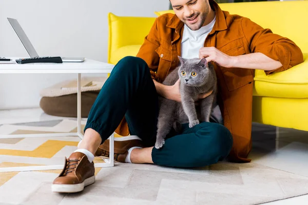 Homem sorridente casual sentado no chão com gato britânico shorthair — Fotografia de Stock