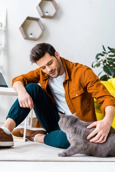 Homem sorridente casual sentado no chão com gato britânico shorthair — Fotografia de Stock