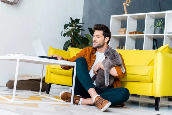 Schöner lächelnder Mann mit Laptop, während er mit britischer Kurzhaarkatze auf dem Boden sitzt — Stockfoto