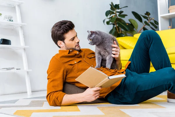 Bell'uomo con libro e sdraiato sul pavimento con simpatico gatto grigio — Foto stock