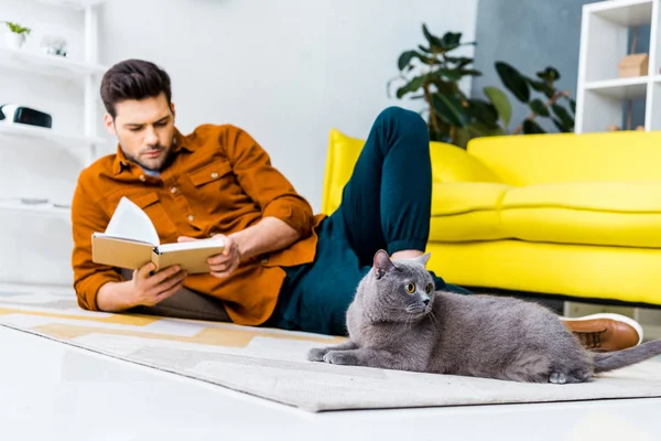 Bell'uomo che legge libro e sdraiato sul pavimento con gatto stenografia inglese in soggiorno — Foto stock