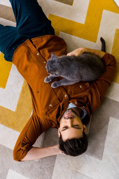 Vista dall'alto dell'uomo pensieroso sdraiato su tappeto con gatto grigio — Foto stock