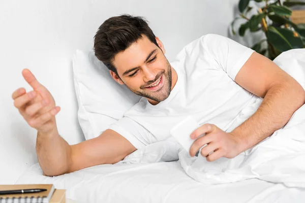 Hombre alegre usando teléfono inteligente en la cama por la mañana - foto de stock
