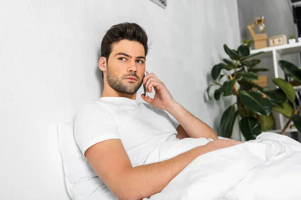 Hombre guapo hablando en el teléfono inteligente mientras descansa en la cama por la mañana - foto de stock