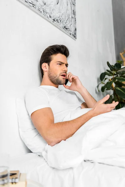 Сердитый человек разговаривает по смартфону, когда отдыхает в постели утром — стоковое фото
