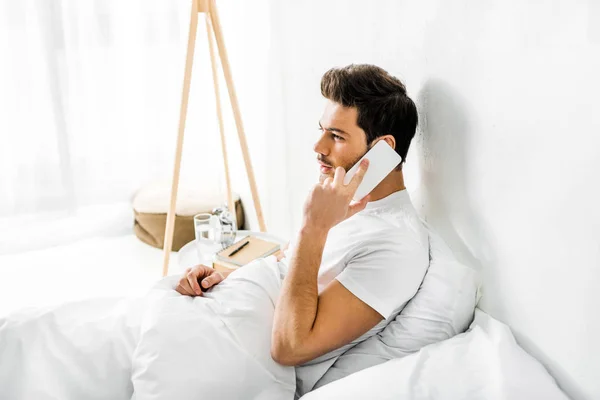 Young man talking on smartphone while sitting on bed in the morning — Stock Photo
