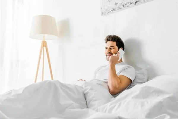 Веселый человек разговаривает на смартфоне, сидя на кровати утром — стоковое фото