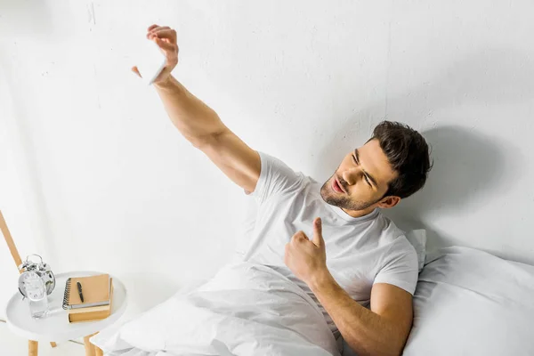 Щасливий чоловік показує великий білий палець, приймаючи селфі на смартфон у спальні — стокове фото