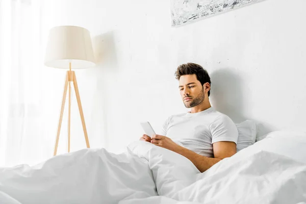 Schöner ernsthafter Mann mit Smartphone, während er morgens im Bett liegt — Stock Photo