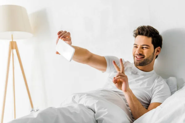 Lächelnder Mann zeigt Friedenssymbol beim Selfie mit Smartphone im Schlafzimmer — Stockfoto
