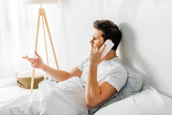 Homem preocupado gesticulando e falando no smartphone enquanto sentado na cama — Fotografia de Stock