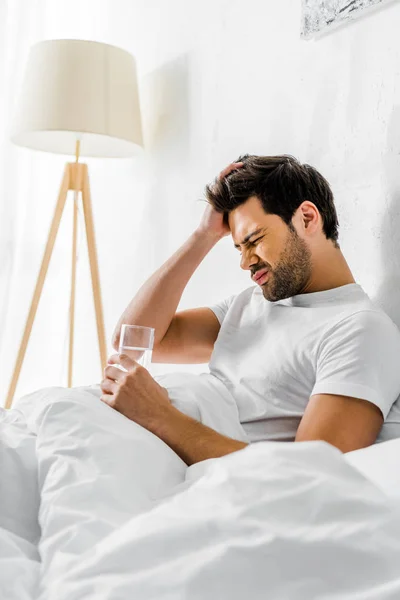 Uomo stanco con postumi di sbornia che tiene un bicchiere d'acqua a letto al mattino — Foto stock