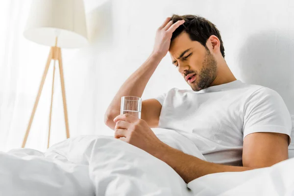 Müder Mann mit Kopfschmerzen, der morgens ein Glas Wasser im Bett hält — Stockfoto