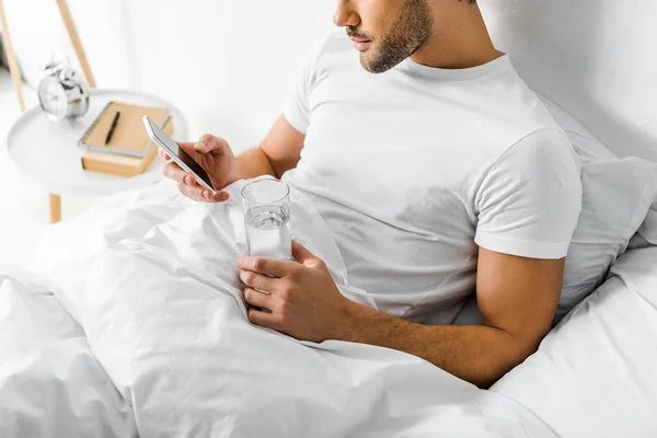 Vista recortada del hombre con vaso de agua usando smartphone en la cama - foto de stock