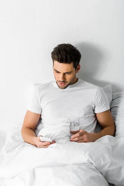 Joven guapo con vaso de agua usando teléfono inteligente en la cama — Stock Photo