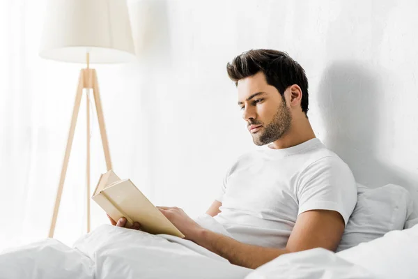 Joven guapo leyendo libro en la cama por la mañana - foto de stock