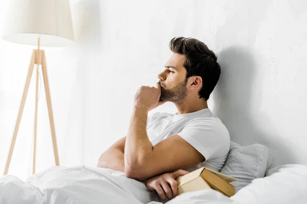 Nachdenklicher junger Mann liegt mit Buch im Bett — Stockfoto