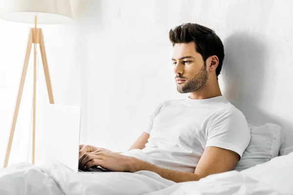 Hombre serio usando el ordenador portátil en la cama por la mañana - foto de stock