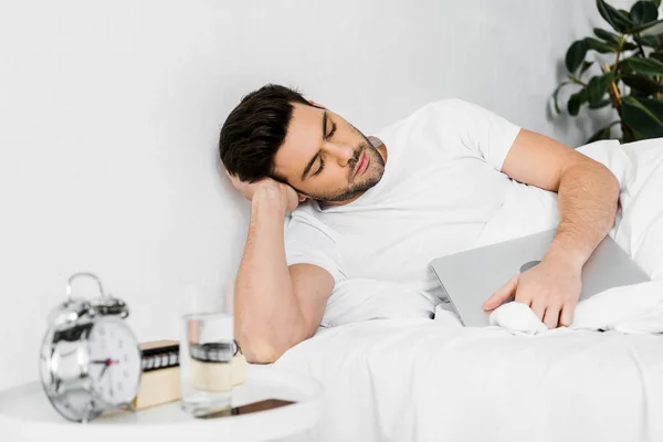 Уставший молодой человек спит с ноутбуком в кровати — стоковое фото