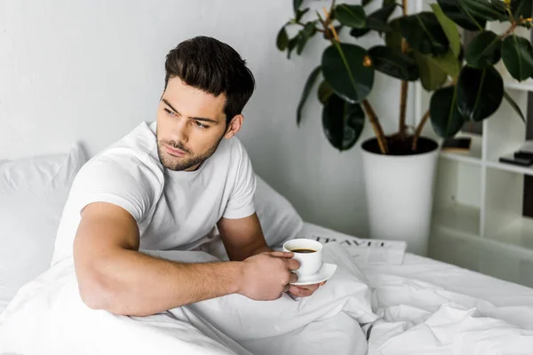 Thoughtful young man in pajamas holding cup of coffee in bed — Stock Photo