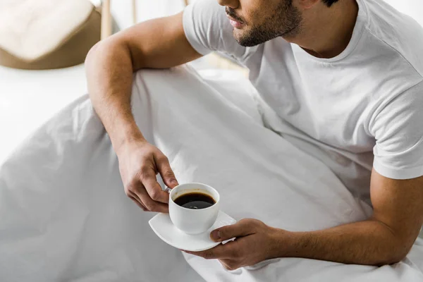 Vista recortada del hombre sosteniendo la taza de café mientras está sentado en la cama - foto de stock