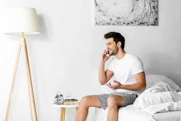 Красивый улыбчивый мужчина, разговаривающий по смартфону, сидя утром на кровати — стоковое фото