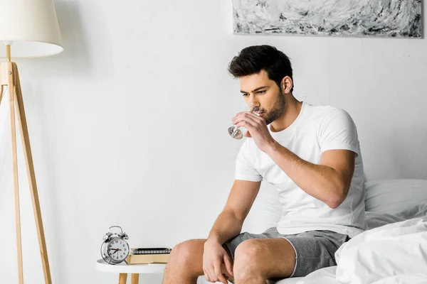 Joven guapo bebiendo agua mientras está sentado en la cama - foto de stock