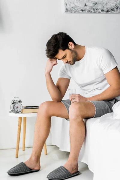 Young man with headache holding glass of water and sitting on bed in the morning — Stock Photo