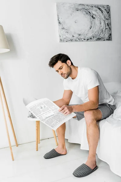 Красивый молодой человек в пижаме читает газету в спальне — стоковое фото