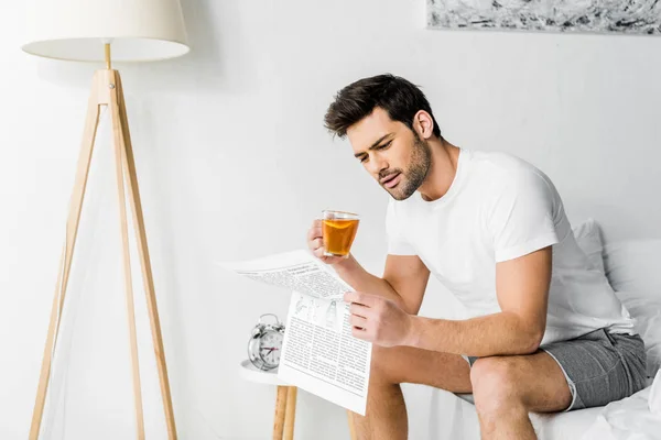 Handsome man with cup of tea reading newspaper in bedroom — Stock Photo